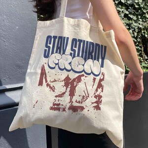May include: Cream-colored tote bag with the words "STAY STURDY CREW" in blue and white bubble letters. Below the text is a maroon graphic of a mountain scene. The bag has two long straps and is made of a canvas-like material.