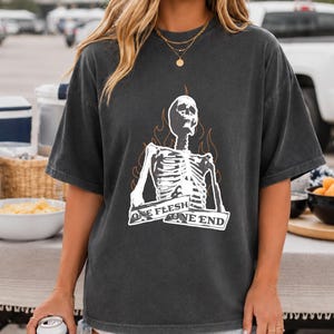 Könnte beinhalten: Ein anthrazitfarbenes Kurzarm-T-Shirt mit einer weißen Grafik eines Skeletts, das von orangefarbenen Flammenkonturen umgeben ist. Ein Banner über der Brust des Skeletts enthält den Text ONE FLESH ONE END in weißer Schrift.