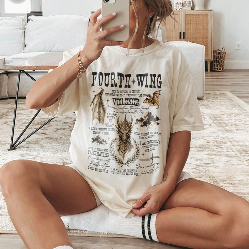 May include: A cream-colored short-sleeve t-shirt featuring a graphic print with the text Fourth Wing and Violence. The design includes illustrations of dragon wings, a central dragon head, a sword, and various lines of small black text.