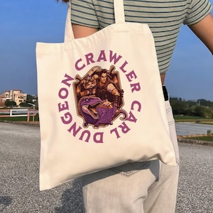 May include: Cream-colored tote bag featuring a circular graphic with the text "Dungeon Crawler Carl" in purple. The graphic depicts a warrior, a cat, and a purple dinosaur. The bag has two long straps.