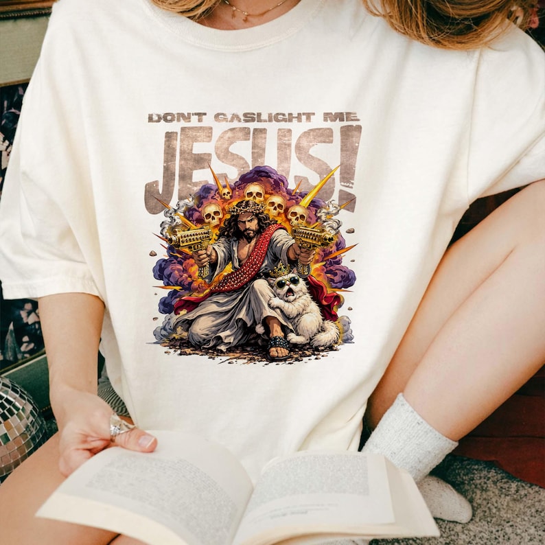 K&ouml;nnte beinhalten: Cremefarbenes T-Shirt mit einer Grafik von Jesus, der zwei goldene Strahlenkanonen h&auml;lt, und einem wei&szlig;en Hund mit Sonnenbrille. Der Text "DON'T GASLIGHT ME JESUS!" ist &uuml;ber dem Bild aufgedruckt. Sch&auml;del und violetter Rauch umgeben das zentrale Bild.
