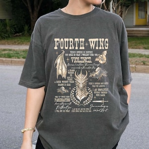 May include: A charcoal gray short-sleeve t-shirt featuring a graphic print with the text "FOURTH WING" and "VIOLENCE." The design includes illustrations of a dragon head, a dragon wing, clouds, a sword, and various quotes in a vintage style.