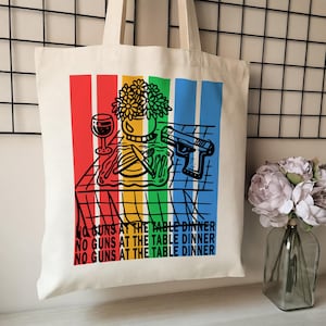 May include: Cream-colored tote bag with a colorful graphic. The design features a wine glass, flowers, a knife and fork, and a handgun. Text on the bag reads "NO GUNS AT THE TABLE DINNER" in red, yellow, green, and blue.