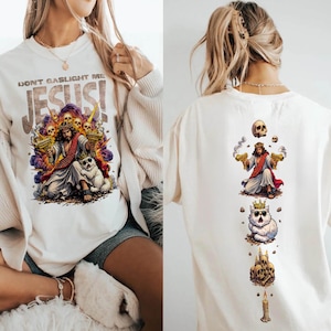 K&ouml;nnte beinhalten: Cremefarbenes Grafik-T-Shirt mit einer farbenfrohen Illustration von Jesus, der zwei goldene Pistolen h&auml;lt, umgeben von Totenk&ouml;pfen und einem Hund. Der Text "DON'T GASLIGHT ME JESUS!" ist &uuml;ber dem Bild aufgedruckt. Die R&uuml;ckseite des Shirts hat ein &auml;hnliches Design.