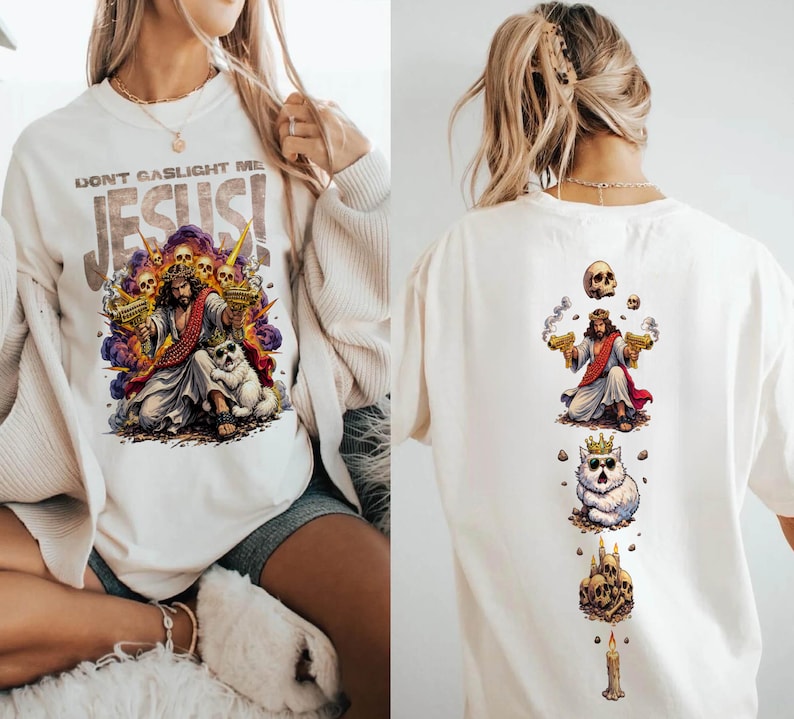 K&ouml;nnte beinhalten: Cremefarbenes Grafik-T-Shirt mit einer farbenfrohen Illustration von Jesus, der zwei goldene Pistolen h&auml;lt, umgeben von Totenk&ouml;pfen und einem Hund. Der Text "DON'T GASLIGHT ME JESUS!" ist &uuml;ber dem Bild aufgedruckt. Die R&uuml;ckseite des Shirts hat ein &auml;hnliches Design.