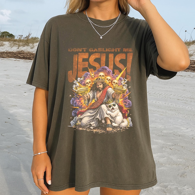K&ouml;nnte beinhalten: Dunkel olivgr&uuml;nes T-Shirt mit einer Grafik von Jesus mit Krone, der zwei goldene Pistolen h&auml;lt, und einem wei&szlig;en Hund mit Krone. Der Text "DON'T GASLIGHT ME" und "JESUS!" ist &uuml;ber der Grafik aufgedruckt.