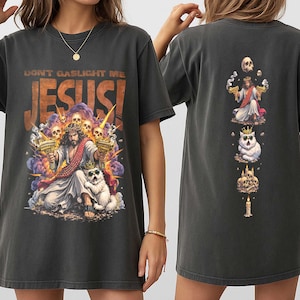 K&ouml;nnte beinhalten: Dunkelgraues T-Shirt mit einem farbenfrohen Grafik von Jesus, der zwei goldene Pistolen h&auml;lt, mit dem Text "DON'T GASLIGHT ME JESUS!". Die R&uuml;ckseite des Shirts hat ein vertikales Design mit Jesus, einer Katze und einer Kerze.