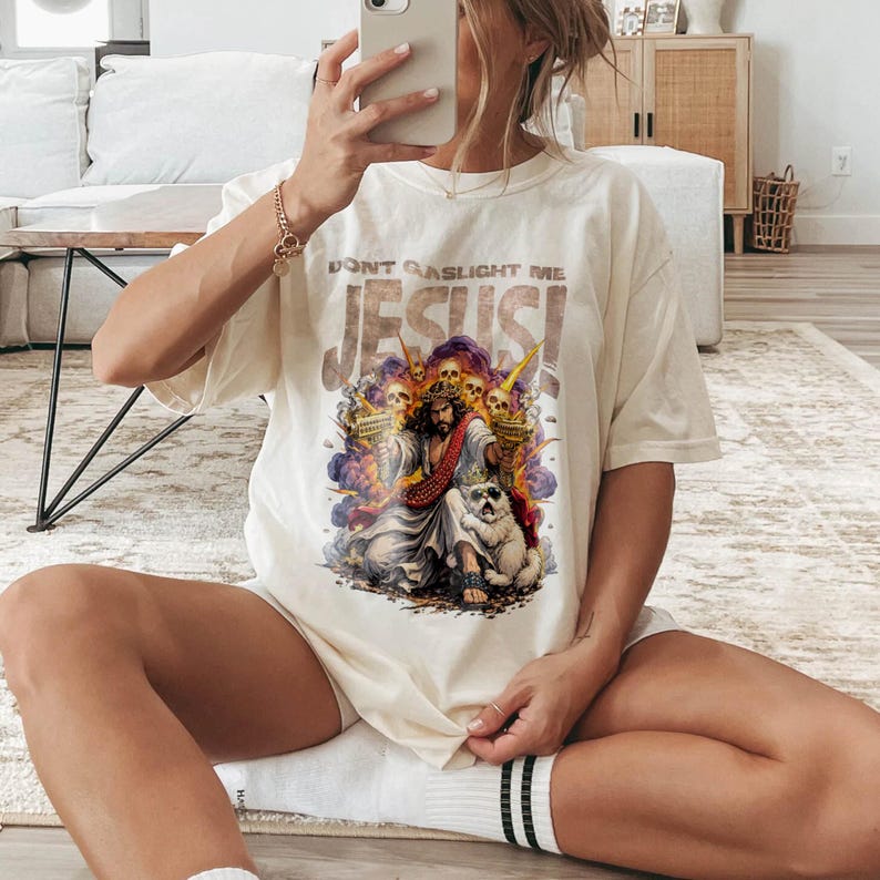 K&ouml;nnte beinhalten: Cremefarbenes T-Shirt mit dem Text "DON'T GASLIGHT ME JESUS!". Die Grafik zeigt eine religi&ouml;se Illustration mit Jesus, einem Hund und Totenk&ouml;pfen. Das Shirt hat kurze &Auml;rmel und eine lockere Passform.