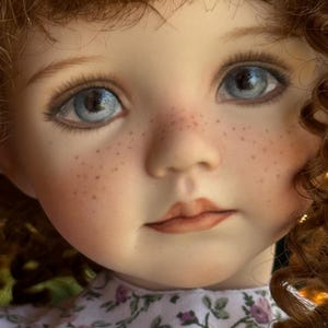 May include: A porcelain doll with curly auburn hair, bright blue eyes, and a dusting of freckles across the nose and cheeks. The doll has a neutral expression with soft pink lips and wears a white garment featuring a small pink and green floral print.
