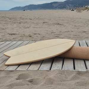 Può includere: Una tavola di equilibrio in legno chiaro dalla forma arrotondata, appoggiata su un rullo di sughero. La tavola ha una sottile linea scura al centro. Gli oggetti sono su una piattaforma di legno vicino a una spiaggia sabbiosa.