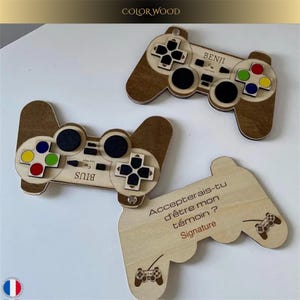 May include: Wooden game controller-shaped favours with engraved details. Two controllers have colourful buttons and the names "BIUS" and "BENJI". A third favour asks "Accepterais-tu d'être mon témoin ?" with a signature line.