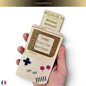 Op de afbeelding: Een houten kaart in Game Boy-vorm met de tekst "THOMAS, VEUX TU ÊTRE MON TÉMOIN?" en "GAME BOY" erop gedrukt. De kaart heeft een sleuf met een kaart met de tekst "GAME NIVEAU SUIVANT". De kaart wordt in een hand gehouden.