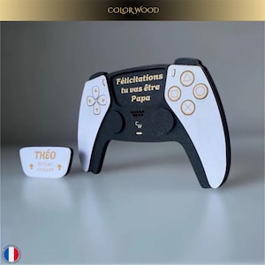Può includere: Un controller in legno bianco e nero con dettagli dorati e la scritta "Félicitations tu vas être Papa". È incluso anche un piccolo pezzo rettangolare con il nome "Théo" e la scritta "Niveau suivant". Tema: videogiochi.