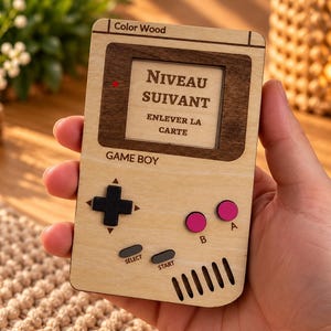 Peut inclure: Une console de jeu portable rectangulaire en bois avec le texte Color Wood, GAME BOY, SELECT, START, A, B et NIVEAU SUIVANT ENLEVER LA CARTE sur l'écran. Elle comprend une croix directionnelle noire, deux boutons roses et des boutons gris.