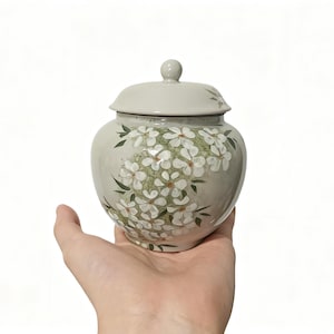 Puede incluir: Un tarro de cerámica con tapa, pintado con flores blancas y hojas verdes sobre un fondo gris claro. El tarro se sostiene en una mano sobre un fondo blanco. Objeto decorativo.