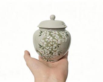 May include: A ceramic jar with a lid, painted with white flowers and green leaves on a light gray background. The jar is held in a hand against a white backdrop. The jar is a decorative item.