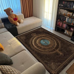 May include: A rectangular brown area rug with a compass design in the center. The rug has a detailed, circular pattern in shades of gold and blue. The rug is placed in a living room with a beige sectional sofa and a bookshelf.