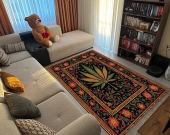 May include: A rectangular area rug with a black background and a large cannabis leaf design in the center. The leaf is yellow and green, surrounded by a floral border in red, orange, and green. The rug is placed on a light-colored floor.