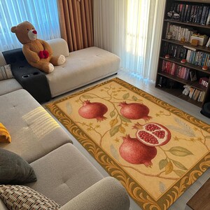 K&ouml;nnte beinhalten: Rechteckiger Teppich mit beigem Hintergrund und Granatapfel-Fruchtmuster. Der Teppich hat einen dekorativen Rand und befindet sich in einem Wohnzimmer mit Sofa und B&uuml;cherregal. Die Ma&szlig;e des Teppichs sind in Zentimetern.