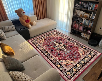 May include: A rectangular red area rug with an ornate floral pattern, bordered by black and cream accents. The rug is placed in a living room setting, with a beige sectional sofa and a bookshelf visible in the background.