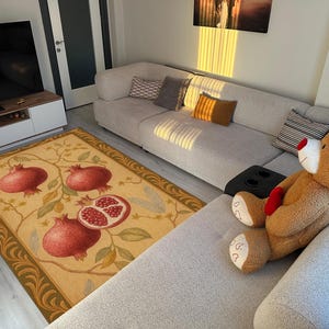 K&ouml;nnte beinhalten: Rechteckiger Teppich mit beigem Hintergrund und gr&uuml;n-goldener Bord&uuml;re. Der Teppich zeigt ein Design aus Granat&auml;pfeln in Rot und Gr&uuml;n. Ein gro&szlig;er Teddyb&auml;r liegt auf einem grauen Sofa.