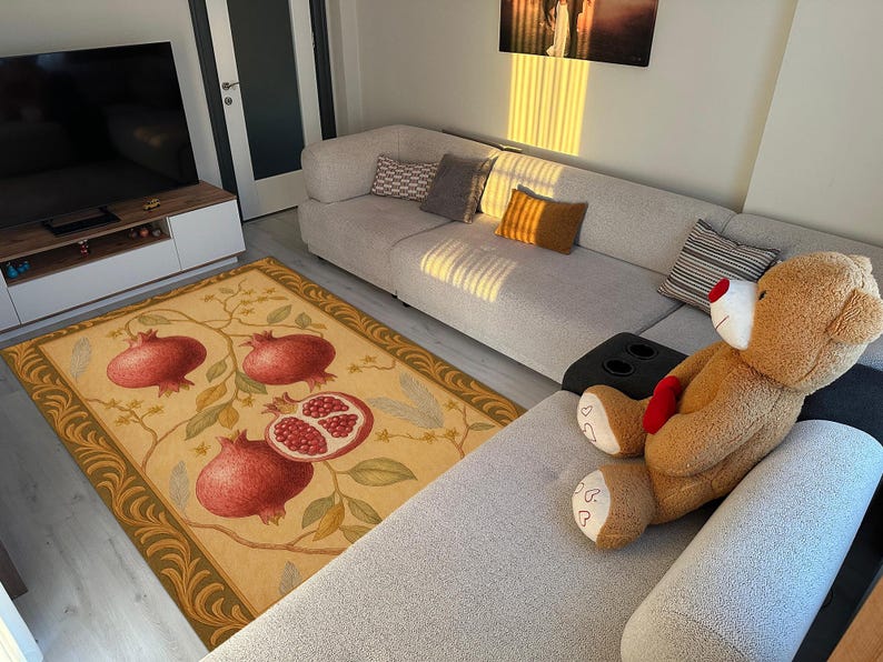 K&ouml;nnte beinhalten: Rechteckiger Teppich mit beigem Hintergrund und gr&uuml;n-goldener Bord&uuml;re. Der Teppich zeigt ein Design aus Granat&auml;pfeln in Rot und Gr&uuml;n. Ein gro&szlig;er Teddyb&auml;r liegt auf einem grauen Sofa.