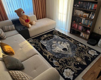 May include: A rectangular black area rug featuring a detailed wolf portrait in shades of gray and white, framed by ornate gold and cream floral designs. The rug is placed on a light-colored floor, with a beige sectional sofa and a bookshelf in the background.