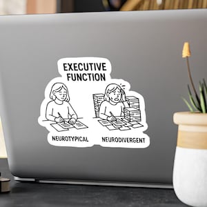 May include: A white sticker on a laptop with the text "EXECUTIVE FUNCTION" above two illustrations. One illustration shows a person writing, labeled "NEUROTYPICAL." The other shows a person overwhelmed by papers, labeled "NEURODIVERGENT."
