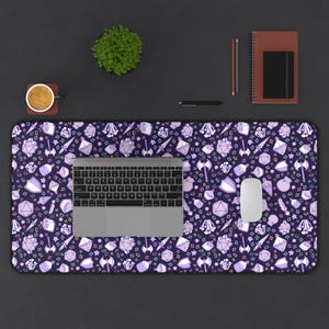 Op de afbeelding: Een paarse bureaumat met een patroon van fantasie-spellen, waaronder dobbelstenen, zwaarden en schilden. Een laptop, muis en andere kantoorbenodigdheden liggen op de mat. Een kopje koffie en een kleine plant zijn ook zichtbaar.