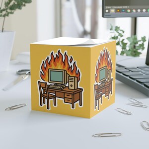 May include: A yellow sticky note cube features a cartoon illustration of a computer desk engulfed in flames. The desk includes a computer monitor, keyboard, CPU, and chair. The design is repeated on multiple sides of the cube.