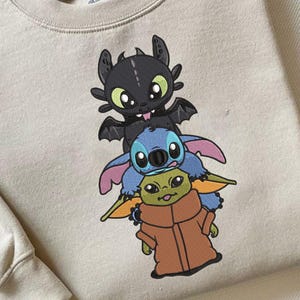 Puede incluir: Sudadera beige de cuello redondo con un diseño bordado de tres personajes de dibujos animados: un dragón negro, un extraterrestre azul y un extraterrestre verde con una túnica marrón. La sudadera tiene cuello y puños acanalados.