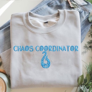 May include: A light gray sweatshirt with the embroidered text "CHAOS COORDINATOR" in blue. Below the text is a blue decorative hook design. The sweatshirt is folded on top of a pair of blue jeans.