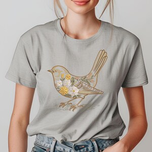 Puede incluir: Camiseta gris claro con una ilustración detallada de un pájaro. El diseño del pájaro es en tonos beige y dorado, con detalles florales en blanco y amarillo. La camiseta tiene mangas cortas.
