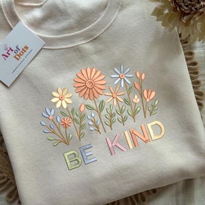 May include: A cream-coloured sweatshirt featuring an embroidered floral design in pastel shades. The design includes various flowers and the words "BE KIND" in a colourful, block letter font. A tag from "Art of Dots" is visible.