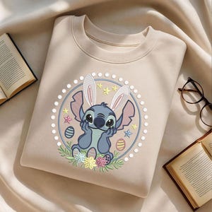 May include: Beige sweatshirt featuring a blue cartoon character with bunny ears, surrounded by Easter eggs and flowers. The design is encircled by a dotted border and stars. A book and glasses are visible in the background.