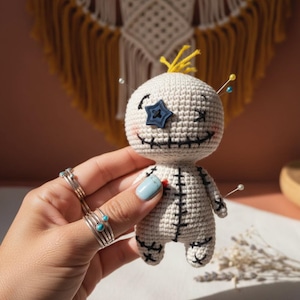 Könnte beinhalten: Handgefertigte, cremefarbene Häkelpuppe mit schwarzen Nahtdetails, einem blauen sternförmigen Knopfauge und gelben Garnhaaren. In die Puppe sind Stecknadeln gesteckt. Die Puppe hat ein lächelndes Gesicht und wird von einer Person mit Ringen an den Fingern gehalten.