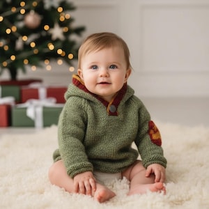 Op de afbeelding: Een baby draagt een groene gebreide hoodie met rode en gele bies en een knoopsluiting. De hoodie heeft een patch op de elleboog. De baby zit op een wit, zacht tapijt voor een kerstboom.