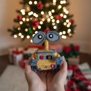 May include: A handmade crochet Wall-E robot toy in yellow, gray, and blue. The toy has large gray eyes and the word "WALL E" is visible. The background features a Christmas tree with blurred lights and wrapped gifts.