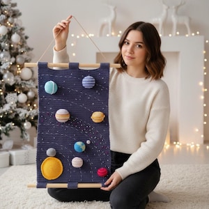 Puede incluir: Un tapiz de pared de ganchillo azul marino con un diseño de sistema solar. Los planetas están representados por bolas de hilo esféricas y coloridas. El tapiz está suspendido de un dowel de madera con una cuerda para colgar.