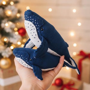 May include: Two handmade crochet whales in navy blue and white. The whales are in a playful pose, with the smaller whale nestled beneath the larger one. The whales have white bellies and dotted details. The background includes a Christmas tree and wrapped gifts.