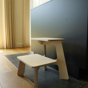 May include: A two-tiered wooden step stool with a natural finish. The stool has a small, square bottom step and a larger, rectangular top step. The design is minimalist and modern, with clean lines and a light wood color.