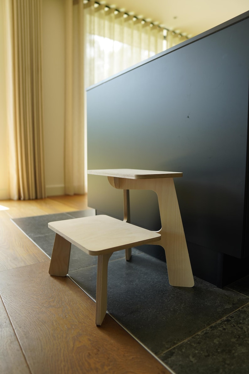 May include: A two-tiered wooden step stool with a natural finish. The stool has a small, square bottom step and a larger, rectangular top step. The design is minimalist and modern, with clean lines and a light wood color.