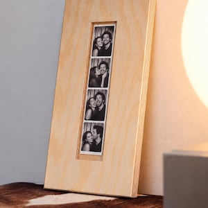 Può includere: Una cornice rettangolare in legno chiaro con un'apertura verticale al centro. All'interno dell'apertura si trova una striscia fotografica in bianco e nero contenente quattro immagini sovrapposte di una coppia.