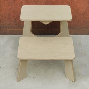 May include: Two wooden step stools, one stacked on top of the other. The stools are made of light-colored wood and have a simple, minimalist design. The top stool has a cutout in the center of the base. The stools are on a concrete surface.