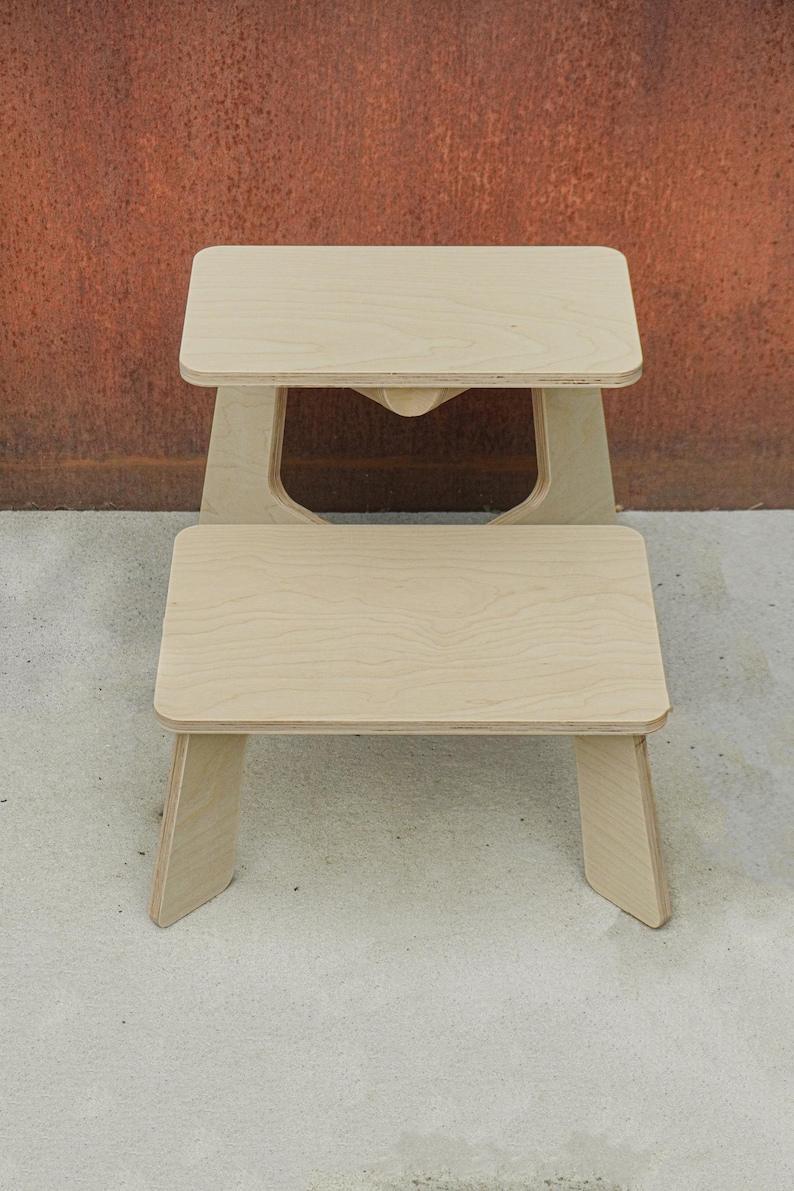 May include: Two wooden step stools, one stacked on top of the other. The stools are made of light-colored wood and have a simple, minimalist design. The top stool has a cutout in the center of the base. The stools are on a concrete surface.