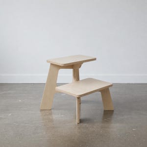 Puede incluir: Un taburete de dos peldaños fabricado en madera contrachapada de color claro con un acabado liso. La estructura cuenta con un peldaño trasero más alto y uno delantero más bajo, ambos apoyados por paneles laterales en ángulo.