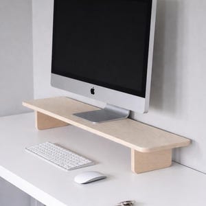 Op de afbeelding: Een lichtgekleurde houten monitorstandaard met een computermonitor erop. Een wit toetsenbord, muis en pen liggen op het witte bureau eronder. De monitor heeft een zwart scherm en een zilveren voet.