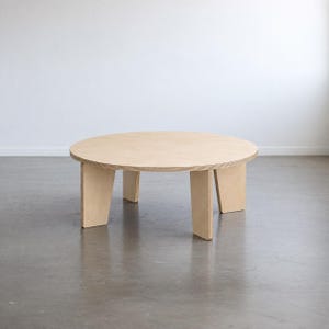 Könnte beinhalten: Ein runder Couchtisch aus hellem Sperrholz. Der Tisch verfügt über eine glatte, kreisförmige Tischplatte und wird von vier markanten, abgewinkelten Beinen getragen, die von der Mitte aus nach außen verlaufen und eine stabile Basis bilden.