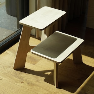 Może przedstawiać: Dwa małe, jasne drewniane stołki. Jeden stołek jest wyższy od drugiego. Stołki mają prostokątne blaty i skośne nogi. Stołki stoją na drewnianej podłodze, z cieniami.