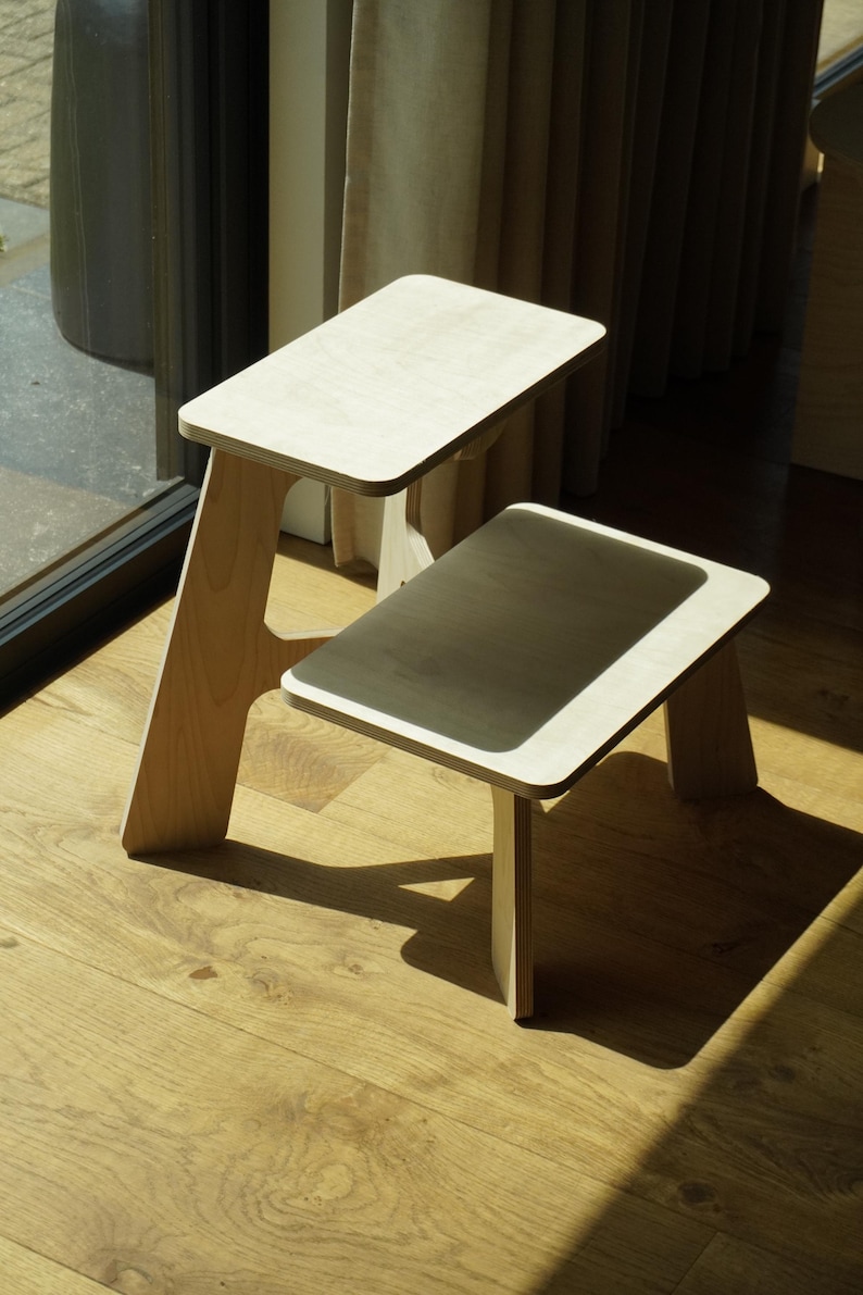 May include: Two small, light-colored wooden stools. One stool is taller than the other. The stools have rectangular tops and angled legs. The stools are on a wooden floor, with sunlight casting shadows.